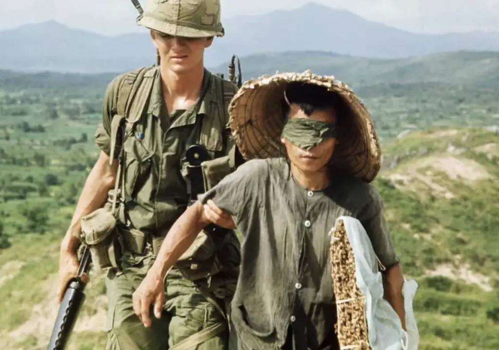 An American soldier is leading a blindfolded Vietnamese individual.