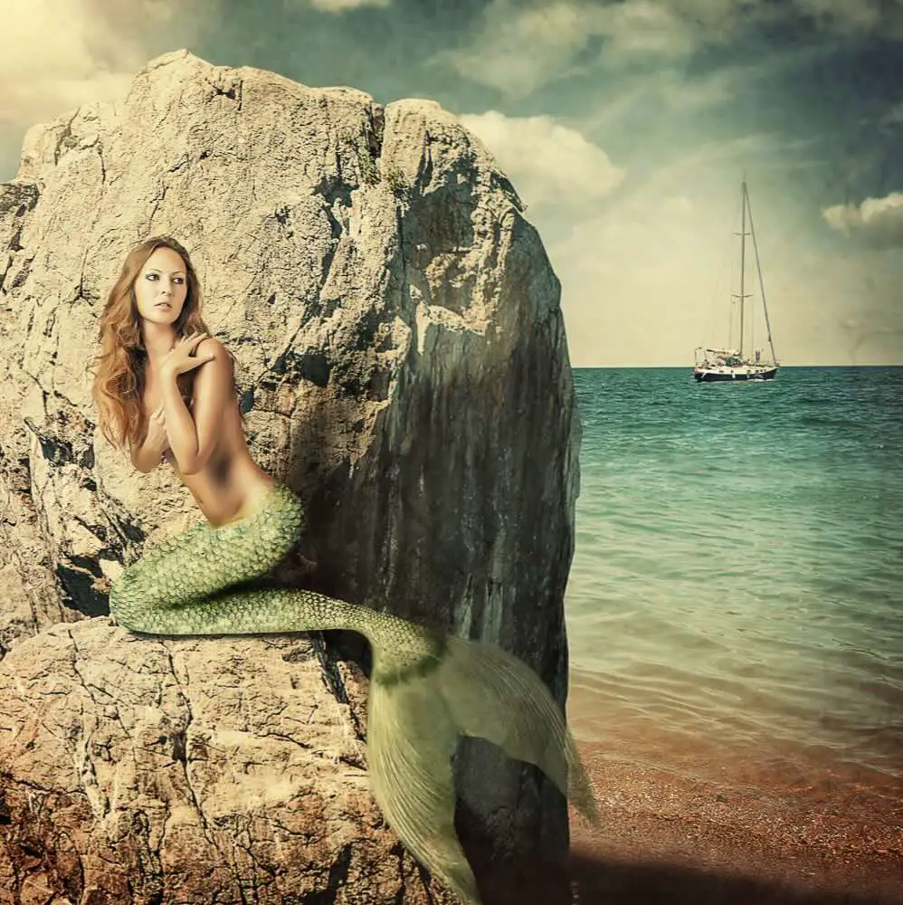 a mermaid sitting on a rock next to a body of water
